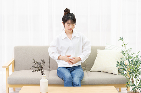 窓際のソファに座ってお腹を手で抑える若い女性